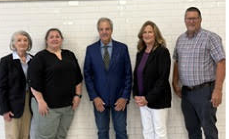A group of five diverse adults stand together indoors against a white tiled wall, smiling for a group photo.